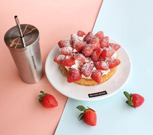 A delicious looking strawberry waffle served on a plate with a stainless steel tumbler beside it at BeansBins Coffee, a popular café in Seoul. Ideal for travelers looking for a must-visit dessert spot.