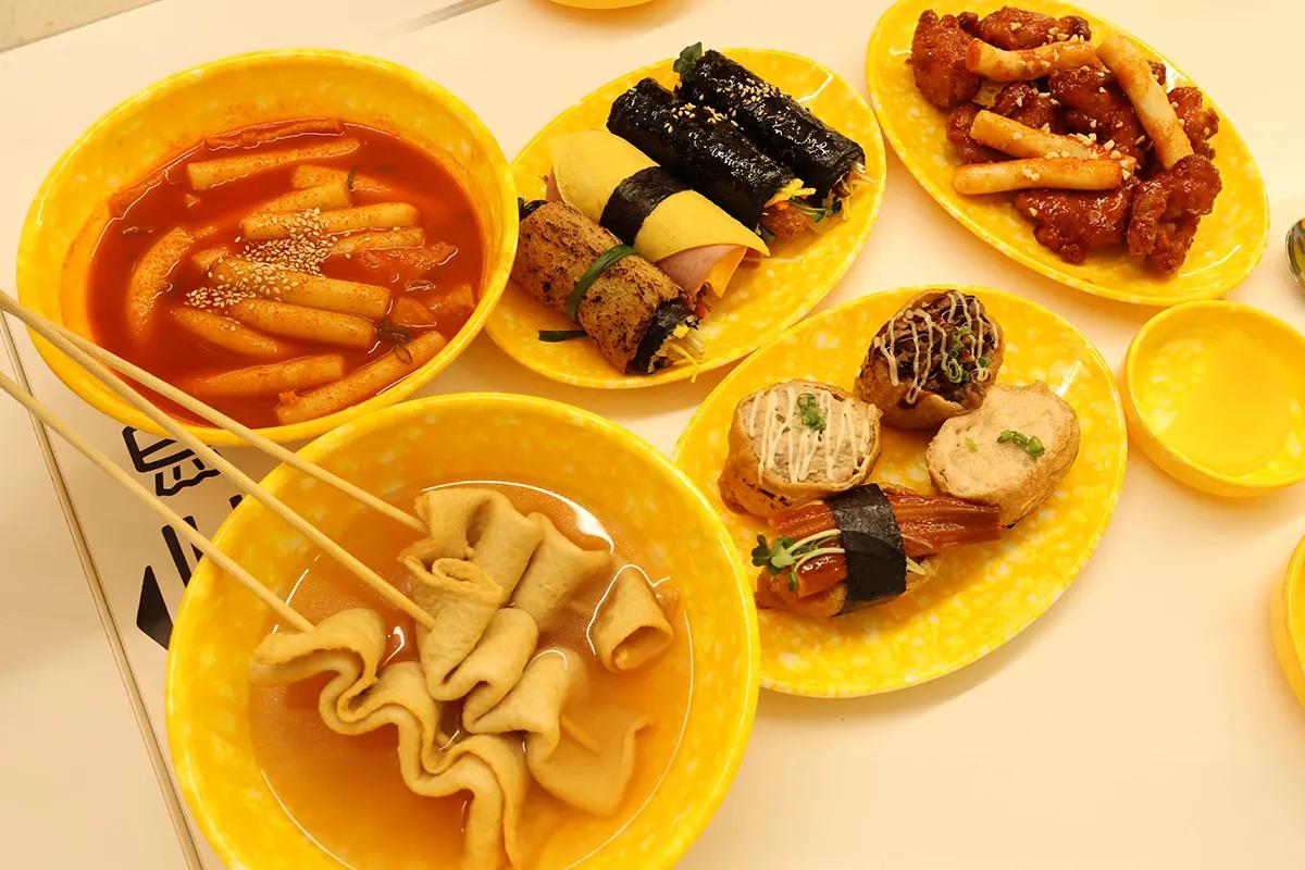 明洞の屋台風店で皿に盛られたトッポッキ、串のオデン、ミニキンパやチキン、揚げ物が並ぶ、手軽にシェアできるひとり飯向けのカジュアルな韓国軽食の盛り合わせ