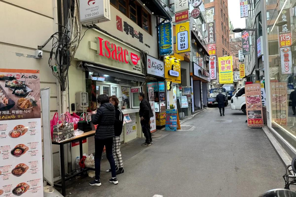 明洞の路地に並ぶテイクアウト店の風景、イサックトーストの屋台看板と並ぶ小さな店と歩道で注文する人々、観光客向けの賑わいを伝える一枚