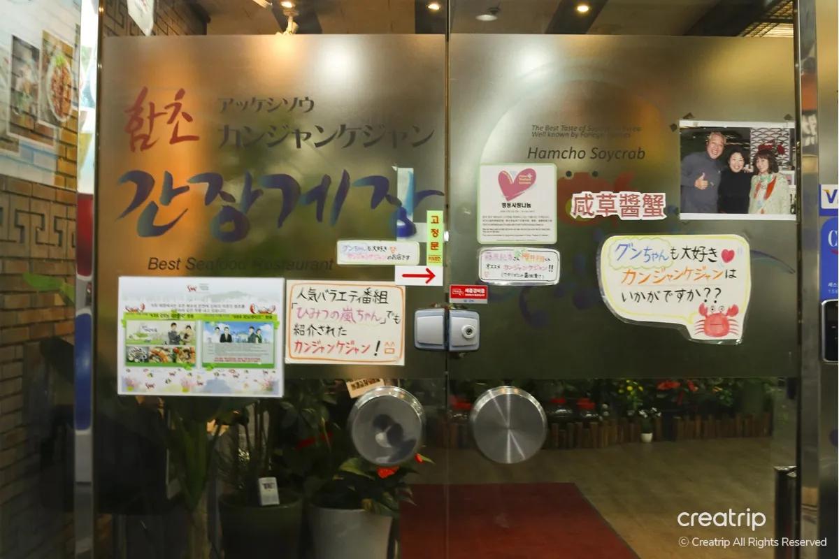 明洞の地下にあるカンジャンケジャン店のガラス扉。店名ロゴや来店写真、メニュー案内が貼られ、日本語の紹介ポップと観葉植物が見える入口の風景。