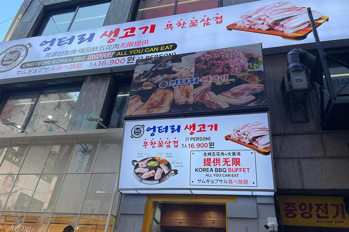 明洞の街並みのビル外壁に並ぶサムギョプサル食べ放題を告知する韓国語・日本語・中国語混在の大型看板と店入口
