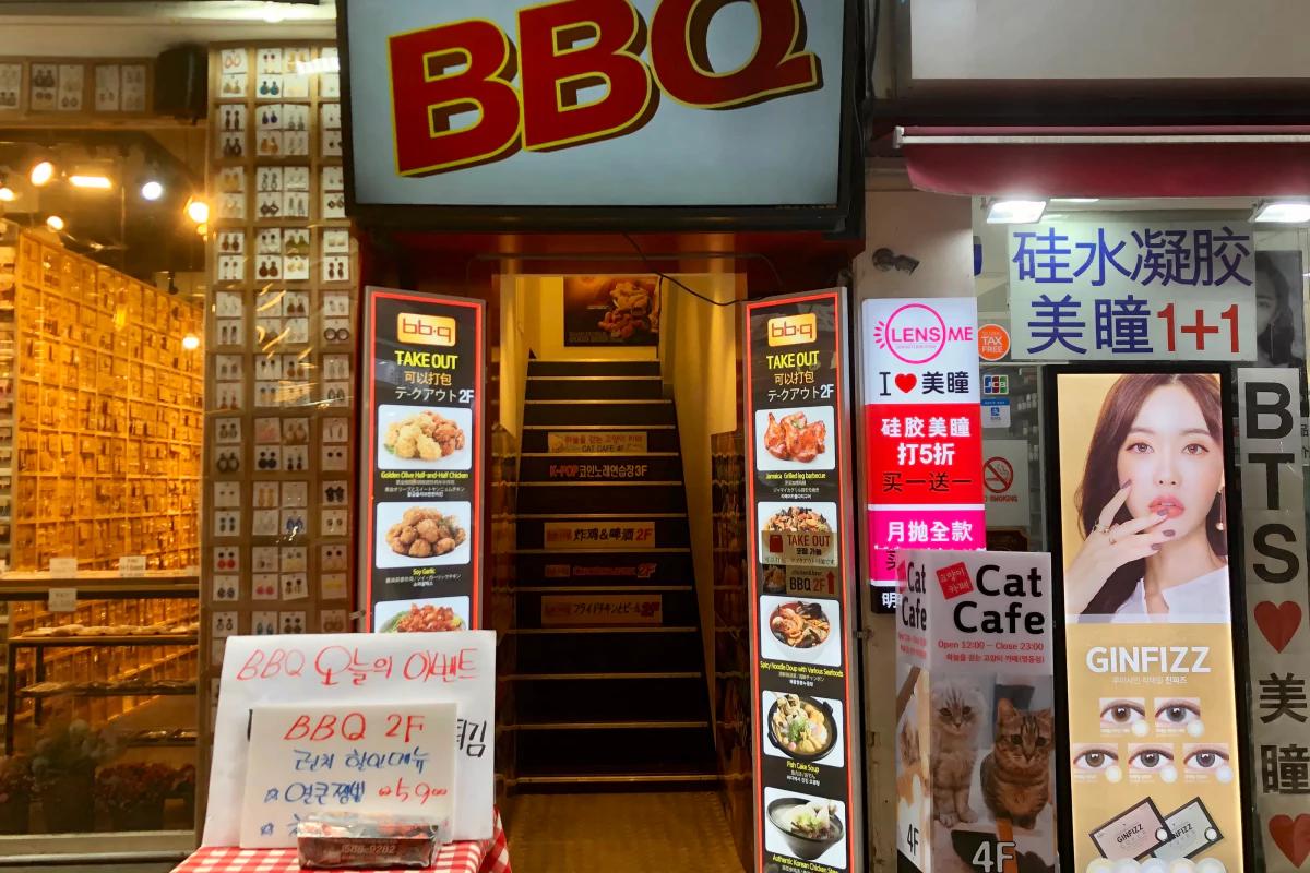 明洞の通りに並ぶ看板と階段、BBQチキンの入口サインと周辺のカフェ・美容ショップの広告が並ぶ夜の風景