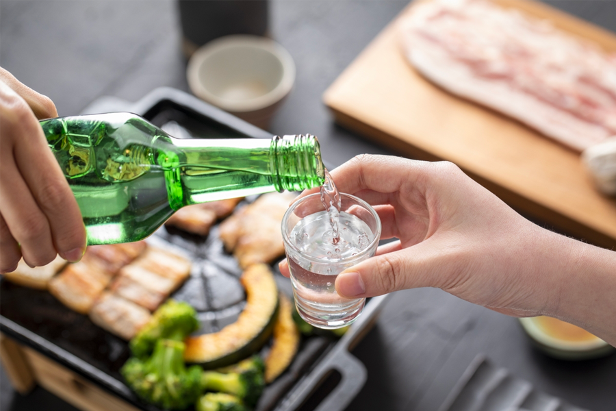 한 손에는 초록색 소주병을 들고, 다른 손에는 작은 잔에 한국 전통 소주를 따르는 모습. 배경에는 삼겹살 구이가 포함된 다양한 야채가 담긴 철판의 이미지.
