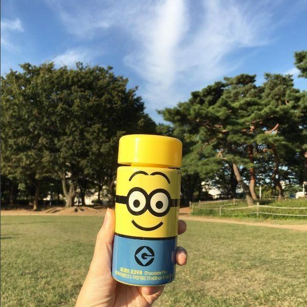 在户外阳光明媚的公园里，小黄人巧克力牛奶瓶拿在手中，背景是一片绿色树林。