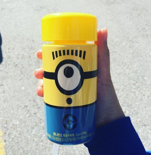 手持小黄人玉米牛奶瓶，背景是模糊的城市街道，瓶身上的小黄人Stuart表情享受。