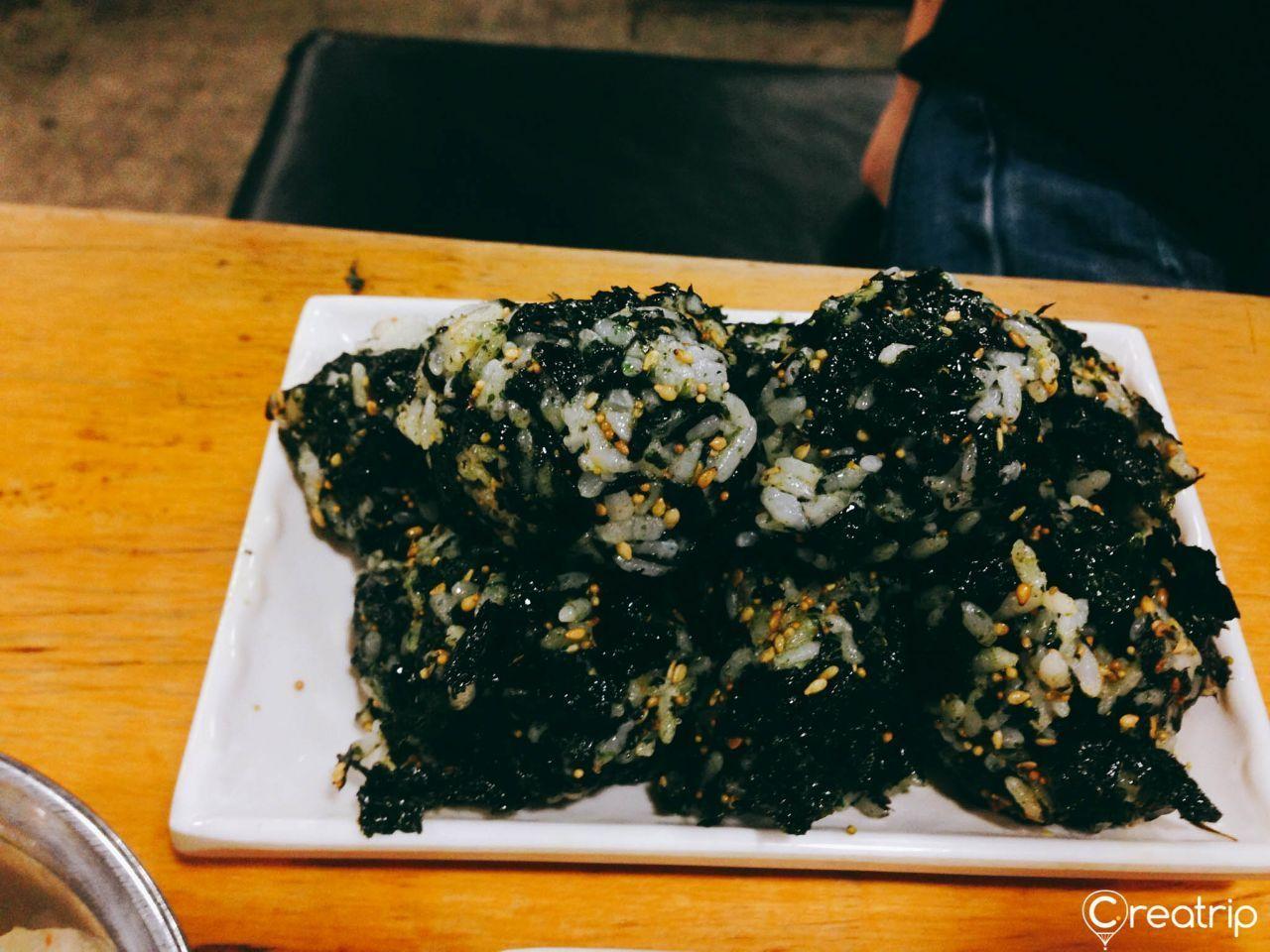 A hand wearing a glove mixing seasoned seaweed with rice to prepare Korean rice balls at Jokbal restaurant.