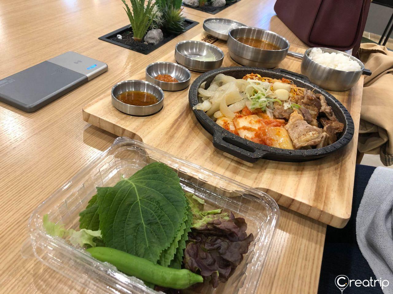 Una mesa servida con una comida de BBQ coreana en un restaurante tipo Yugcheobbansang, donde se puede ver una variedad de platos incluyendo kimchi, carne a la parrilla, y una bandeja de vegetales como lechuga y hojas de perilla, dentro de un ambiente moderno y acogedor con utensilios de acero inoxidable.