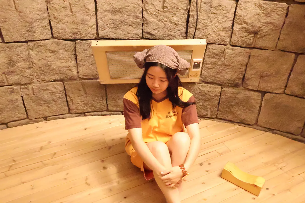 穿着傳統韓服坐在桑拿房內的女人,頭上用毛巾做成羊角造型,背景是石牆。