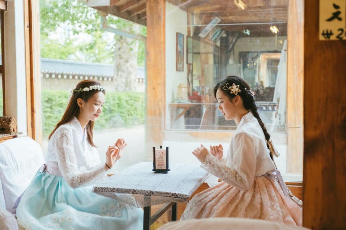 Two women sharing a moment inside a quaint traditional Korean house, both wearing pastel Hanboks and floral crowns.