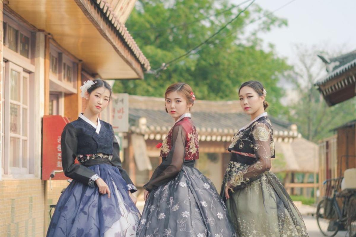 Three women elegantly posed in vibrantly colored Hanboks outside a traditional Korean village building, with indicative floral patterns on their dresses.