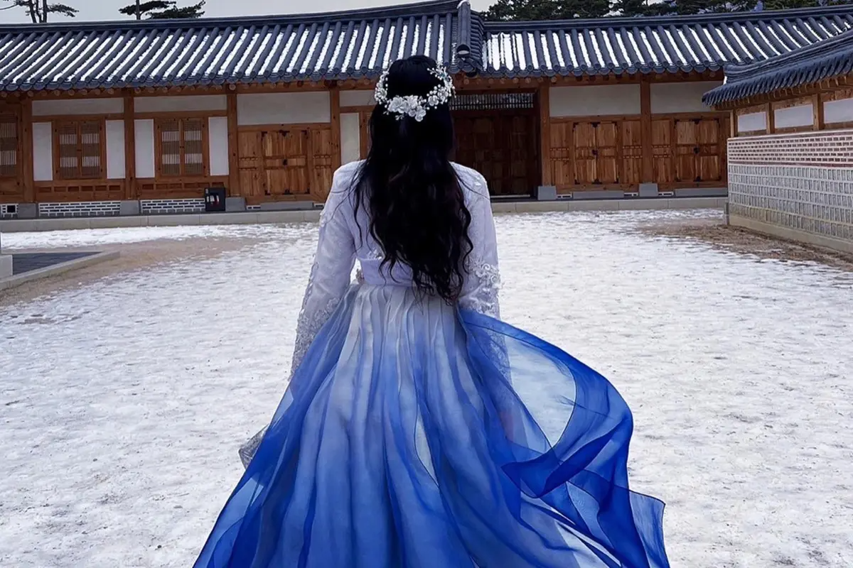 A woman in an elegant blue Queen Hanbok walking away from the camera towards a traditional Korean building. Her Hanbok features a flowing blue skirt with a floral hairpiece.