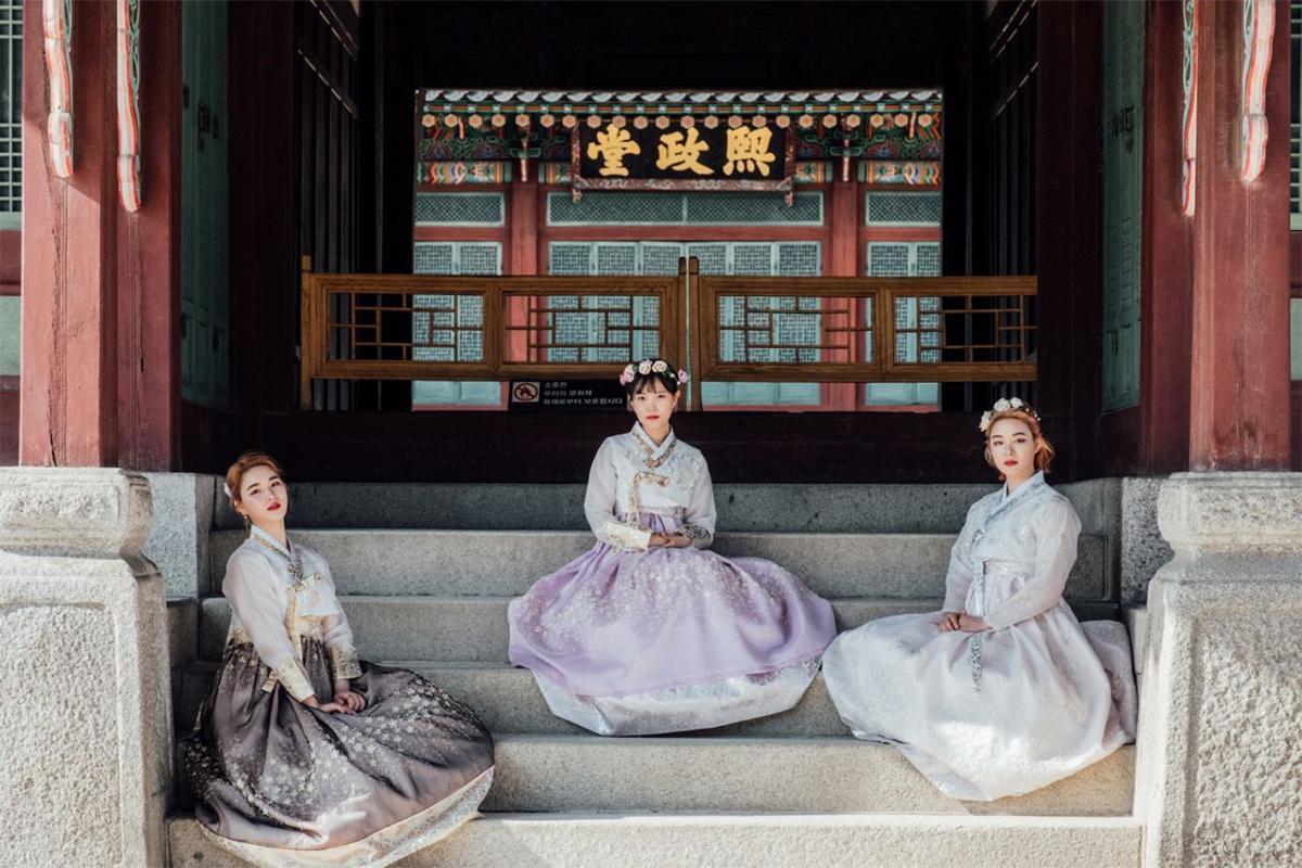 Three women dressed in pastel Hanboks sitting gracefully on the steps inside a traditional Korean palace, decorated with ornate hairpieces.
