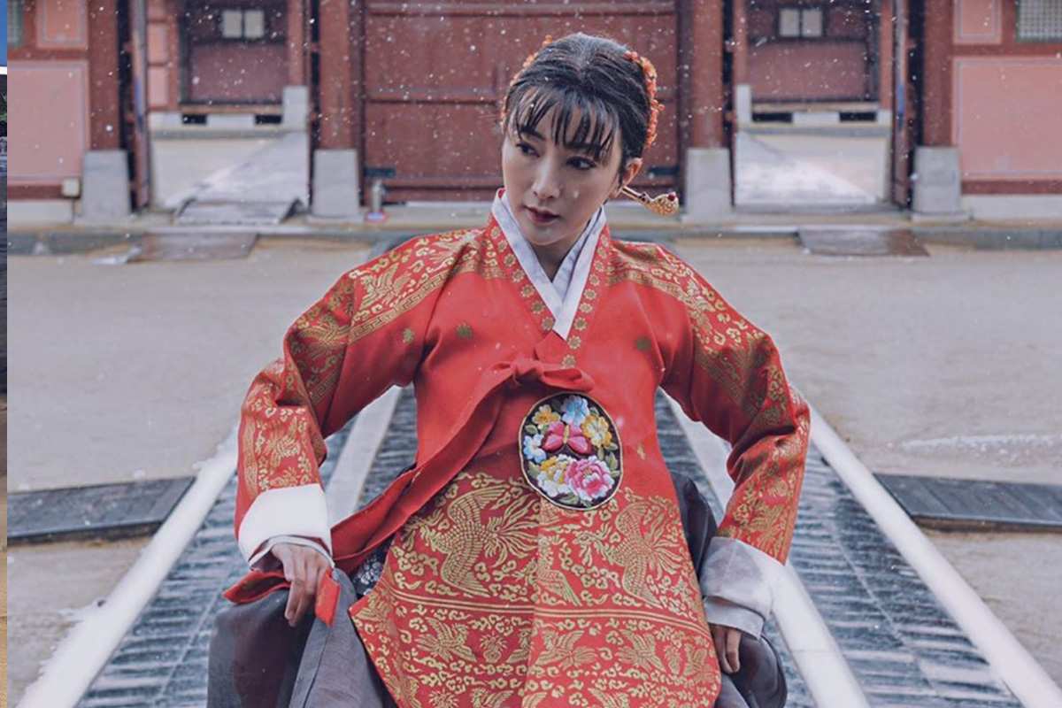 一名身穿紅色王妃韓服的女性在雪中於首爾韓國傳統建築前跪坐,韓服上的金色刺繡細節凸顯出韓式服飾的奢華與傳統。」