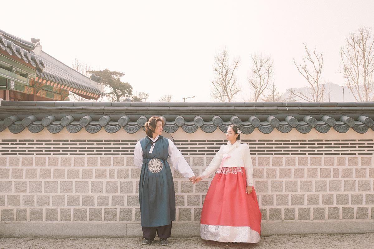 A couple holding hands while posing next to a traditional Korean wall, dressed in complementary Hanbok outfits, his in blue and hers in red.