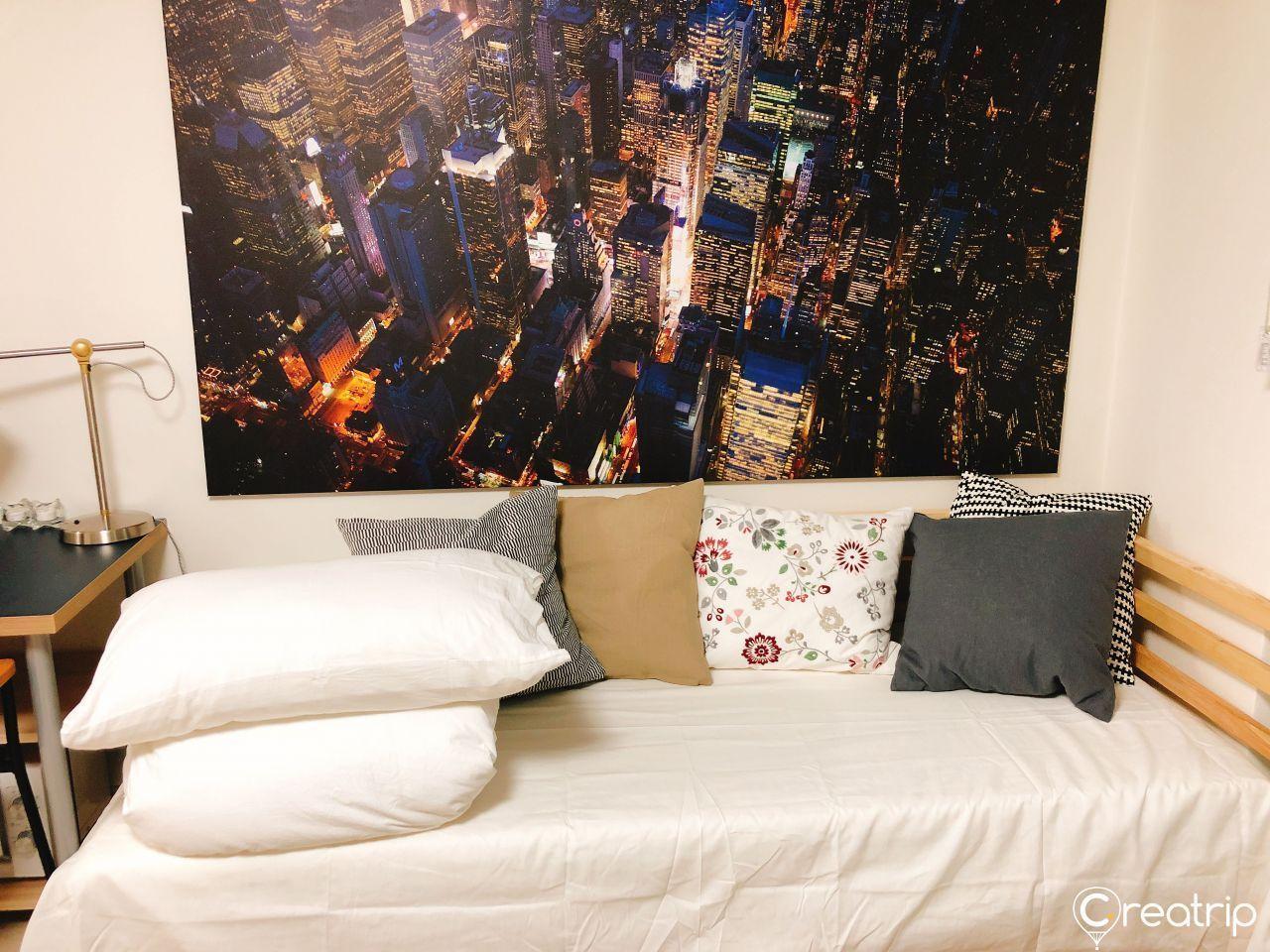 一間簡約明亮的卧室，單人牀上擺放著多個枕頭，牆上掛著一幅夜景城市畫像。