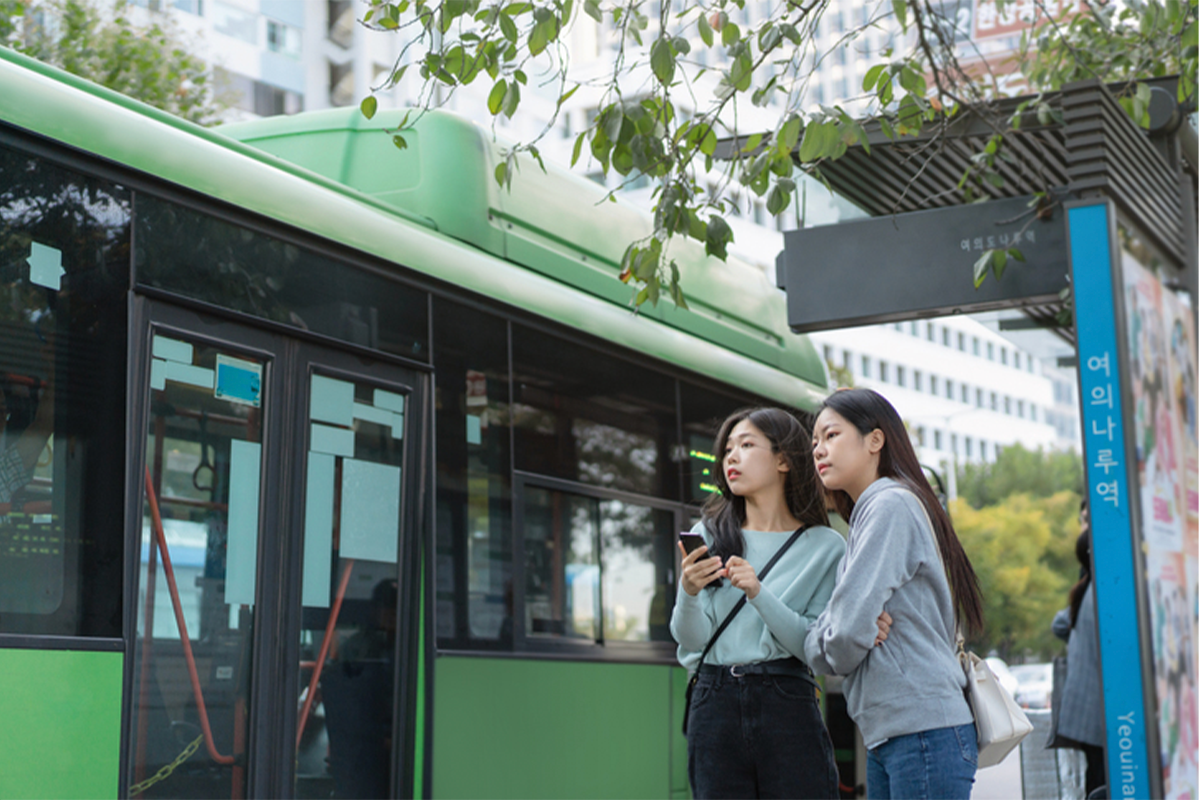 綠色公共汽車旁的路邊，兩位女士正在查看手機，體現韓國的公共交通便利。