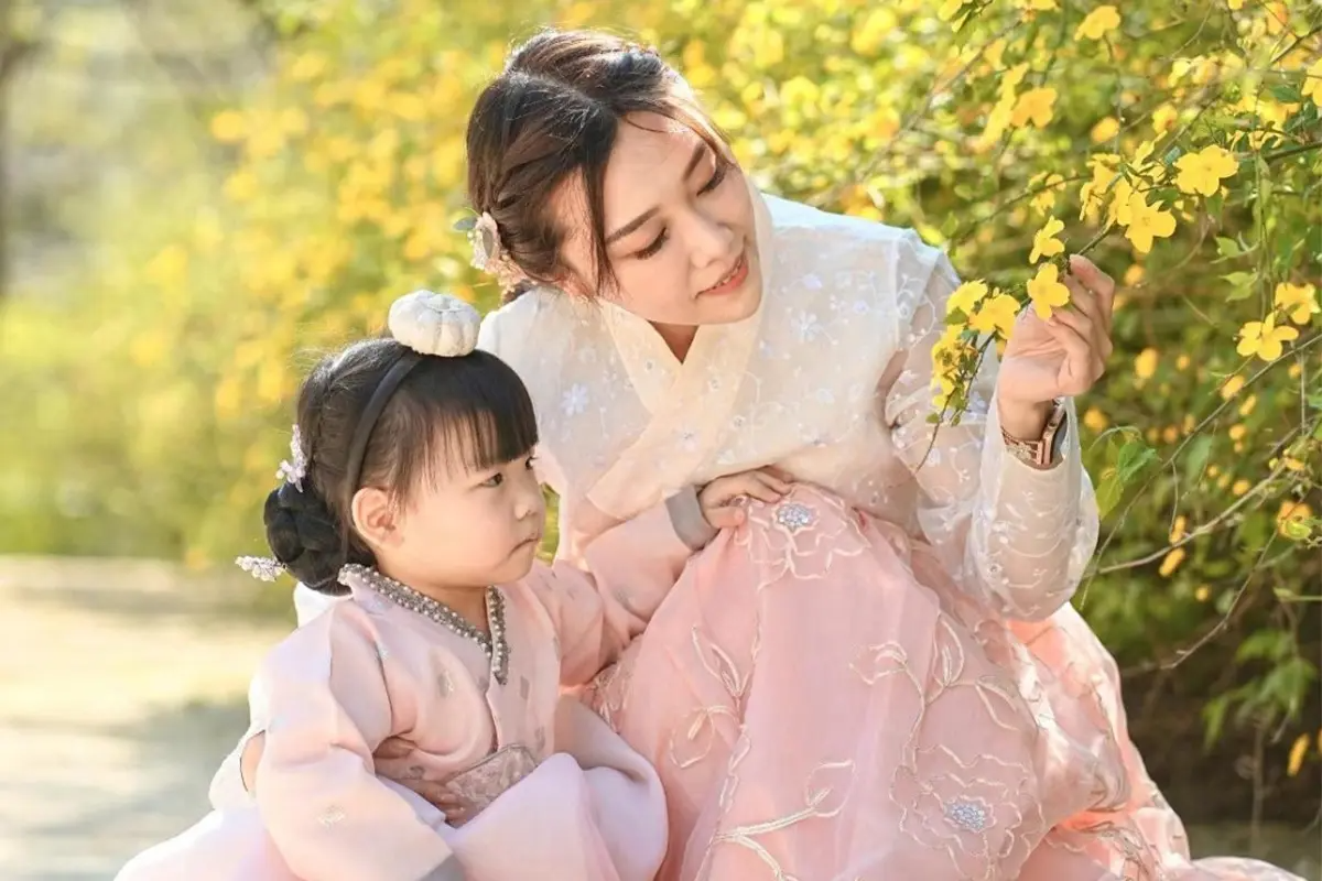 一位母親與她的小女兒穿著粉色小童韓服,背景是盛開的黃色花叢。