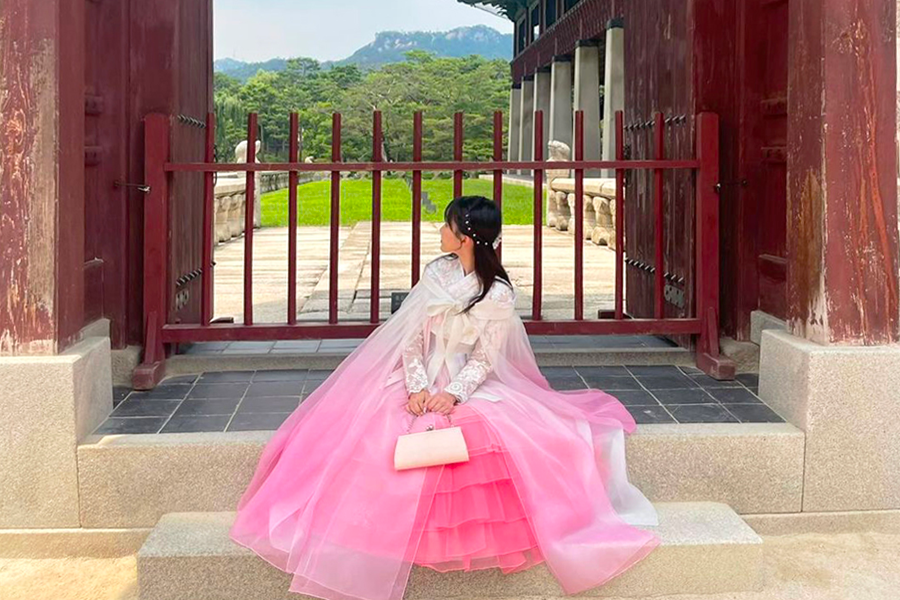 Woman wearing a pink hanbok sitting at a traditional Korean gate, looking into the distance.