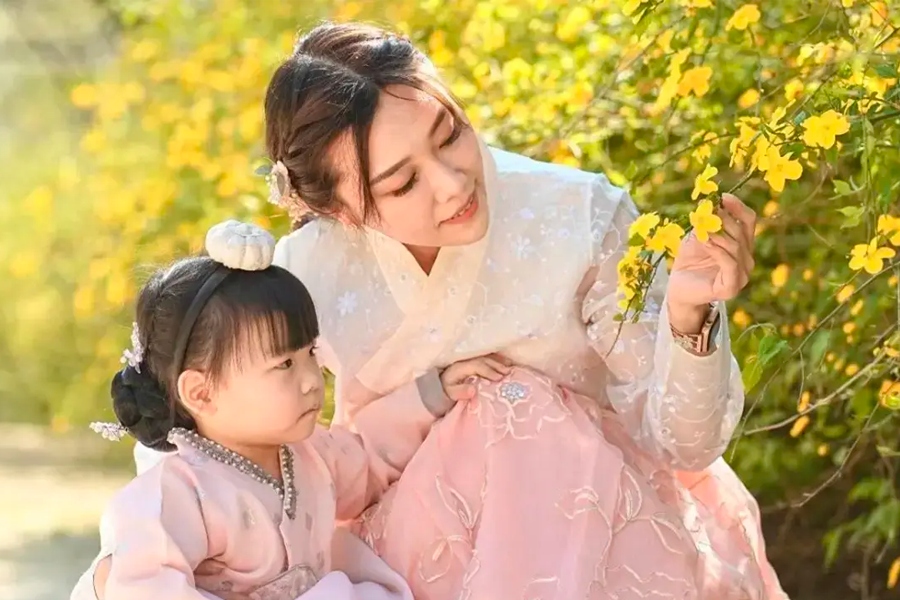 Mother and child wearing pastel-colored hanboks with floral details, sitting amidst yellow flowers.