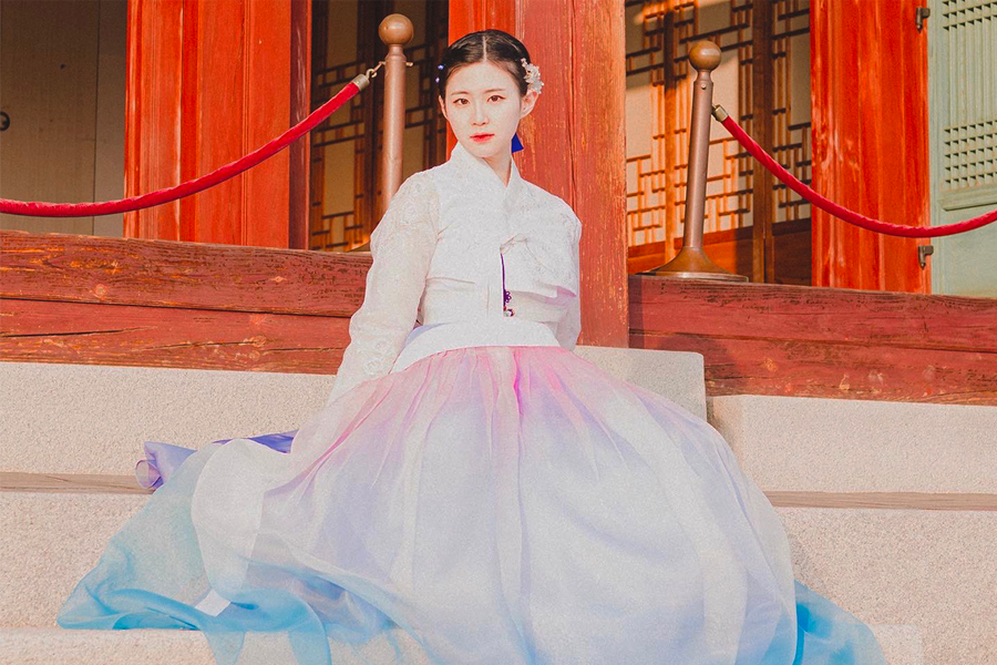 Person wearing a hanbok seated on steps in front of a traditional Korean building.