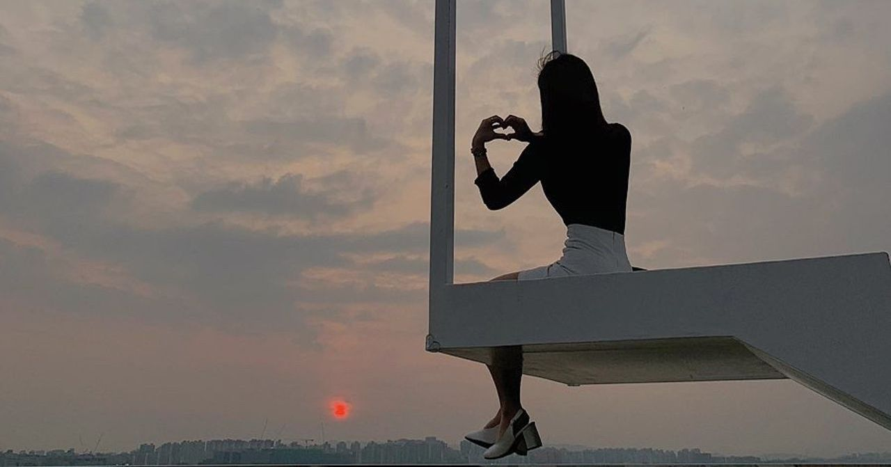 一位女性坐在日落的天空階梯上，雙手擺成心型，穿著黑色上衣和白色裙子，背景是壯觀的城市天際線，捕捉寧靜和夢幻的奇蹟時刻。