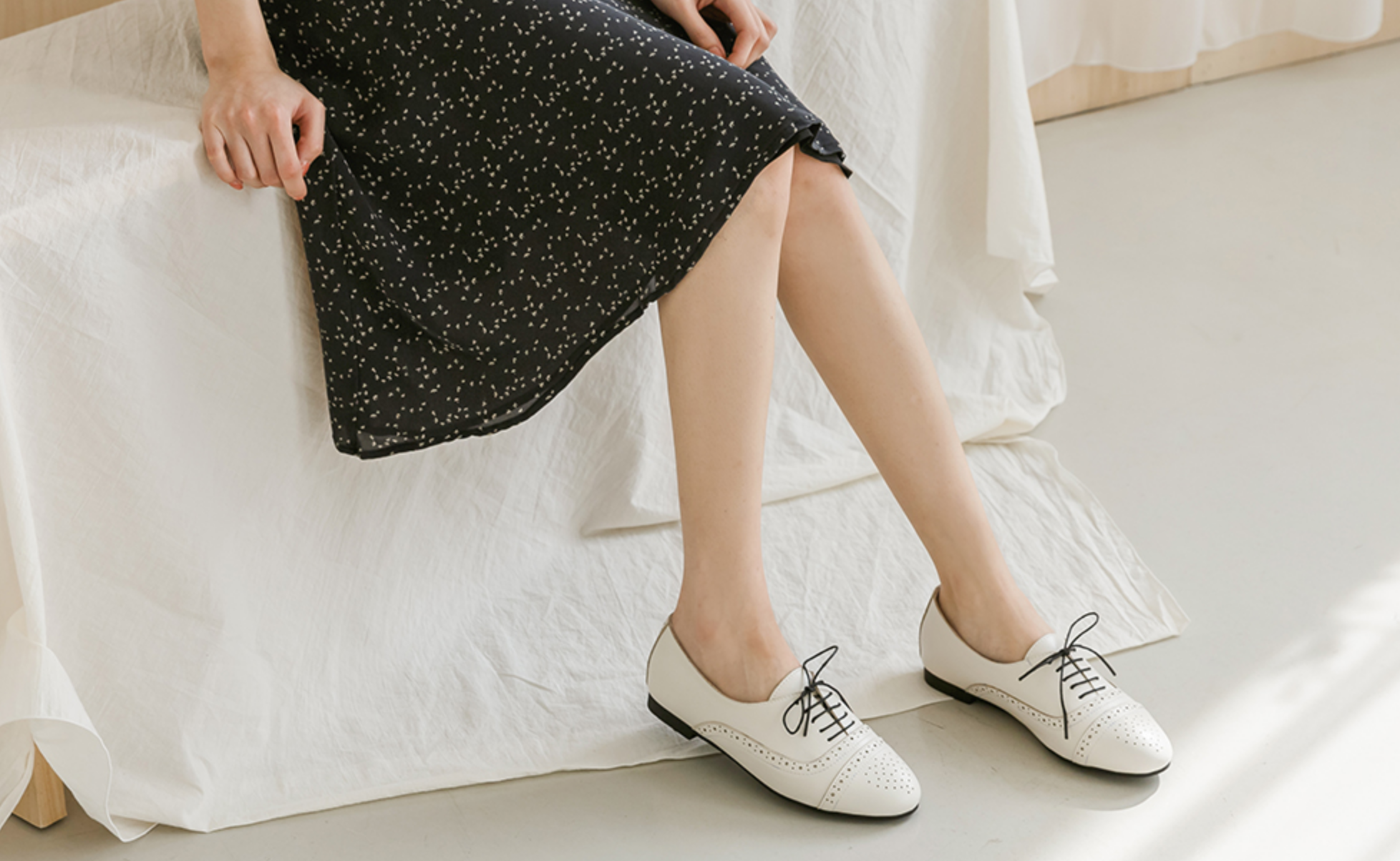Woman sitting with crossed legs wearing white lace-up shoes and a black dress, highlighting the elegant design of BFSOLE footwear.