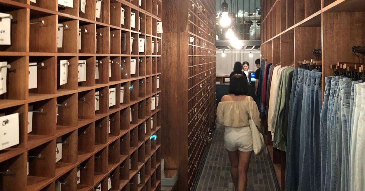 Wide hallway with wooden shelving units filled with small boxes and hangers of denim, possibly in a fashion retail space.