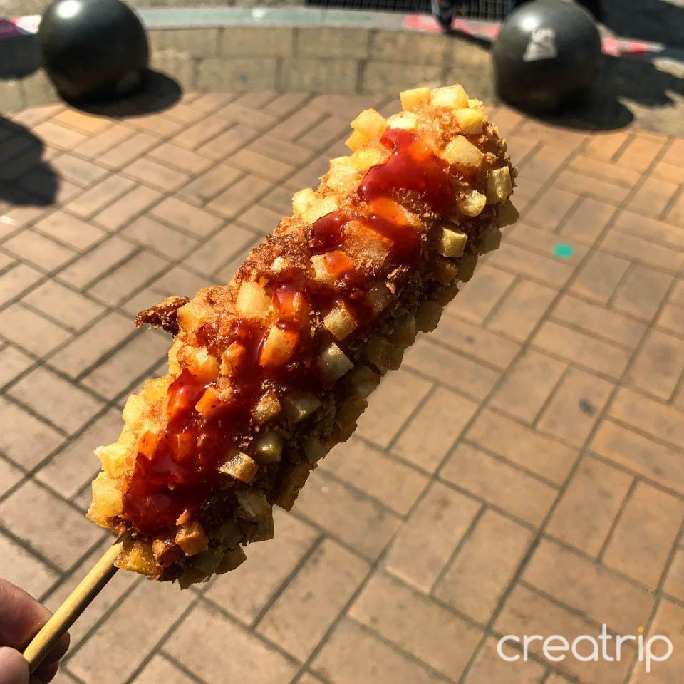 Close-up of Korean legendary street treat '熱狗' covered in golden-brown batter and crispy diced potatoes, captured perfectly with a squeeze of ketchup.