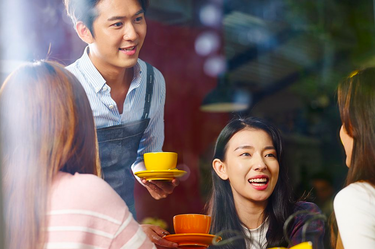 一位咖啡館服務員正在與顧客交流並遞送咖啡。畫面中顯示著歡樂的聚會和輕鬆的咖啡館環境。