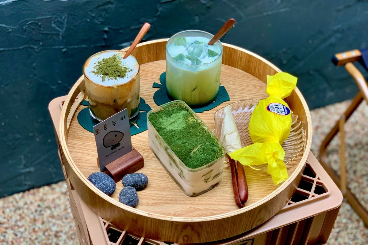 木製託盤上擺放著兩杯抹茶拿鐵和一份抹茶提拉米蘇甜點，旁邊有木製餐具和裝飾物，展現精緻的咖啡館飲品與甜點。