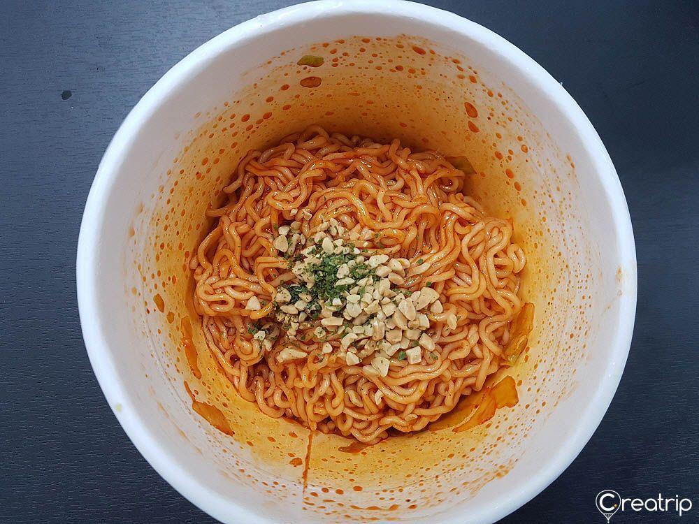 Cooked spicy and sweet chicken flavor instant noodles in a bowl, topped with seasoning and nuts