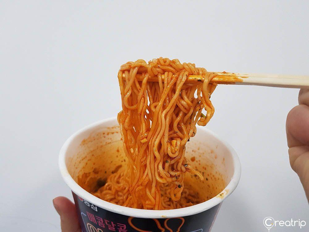 杯裝即食麵條從白色紙杯裡托起,辣醬紅色且吸引眼球,麵條緊密纏繞在筷子上,出現在手中,背景為白色,展現辣味與韓國便利店人氣商品的獨特色彩。