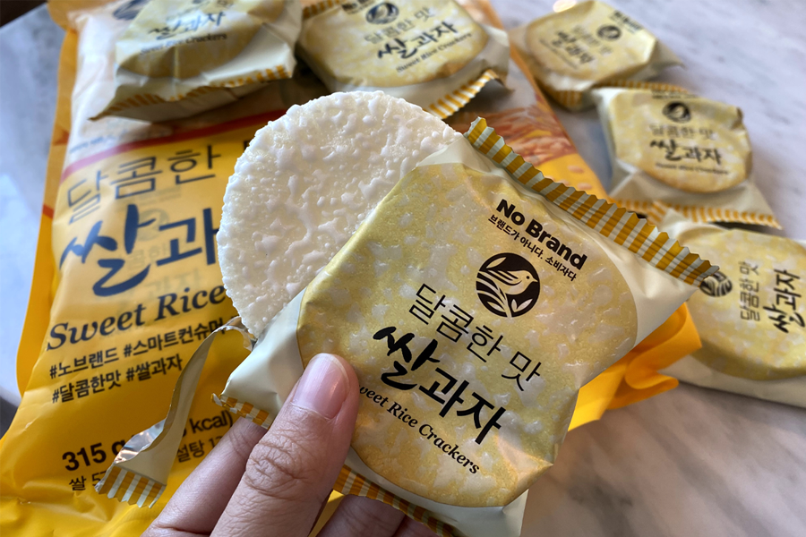 A hand holding a rice cracker from a packed yellow bag of No Brand's sweet rice crackers, showcasing their light and crispy texture ideal for a guilt-free snacking option.