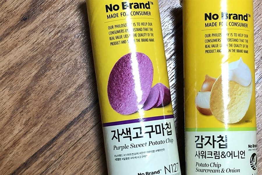 Two No Brand cylindrical canisters on a wooden surface, one featuring purple sweet potato chips and the other sour cream and onion potato chips. The vibrant colors and unique flavors make them stand out as a must-try Korean snack.