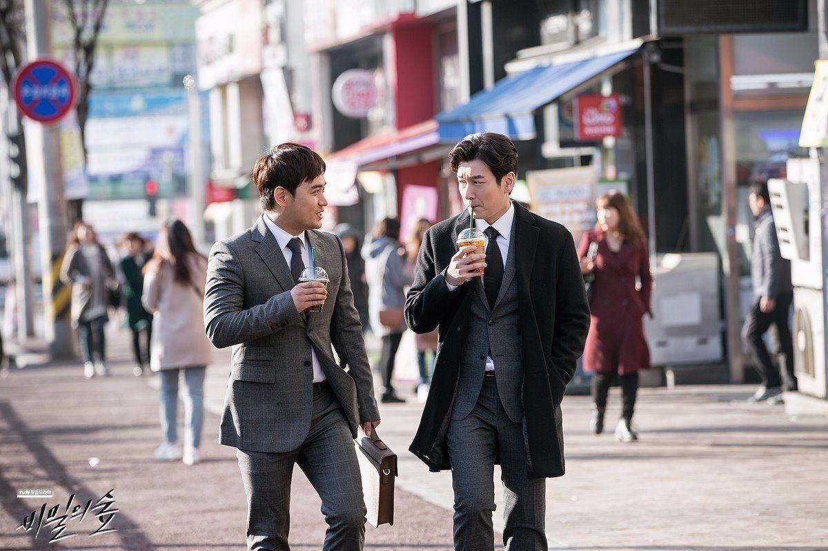 两位穿西装的男性在城市街道中步行，每人手中持有饮料。