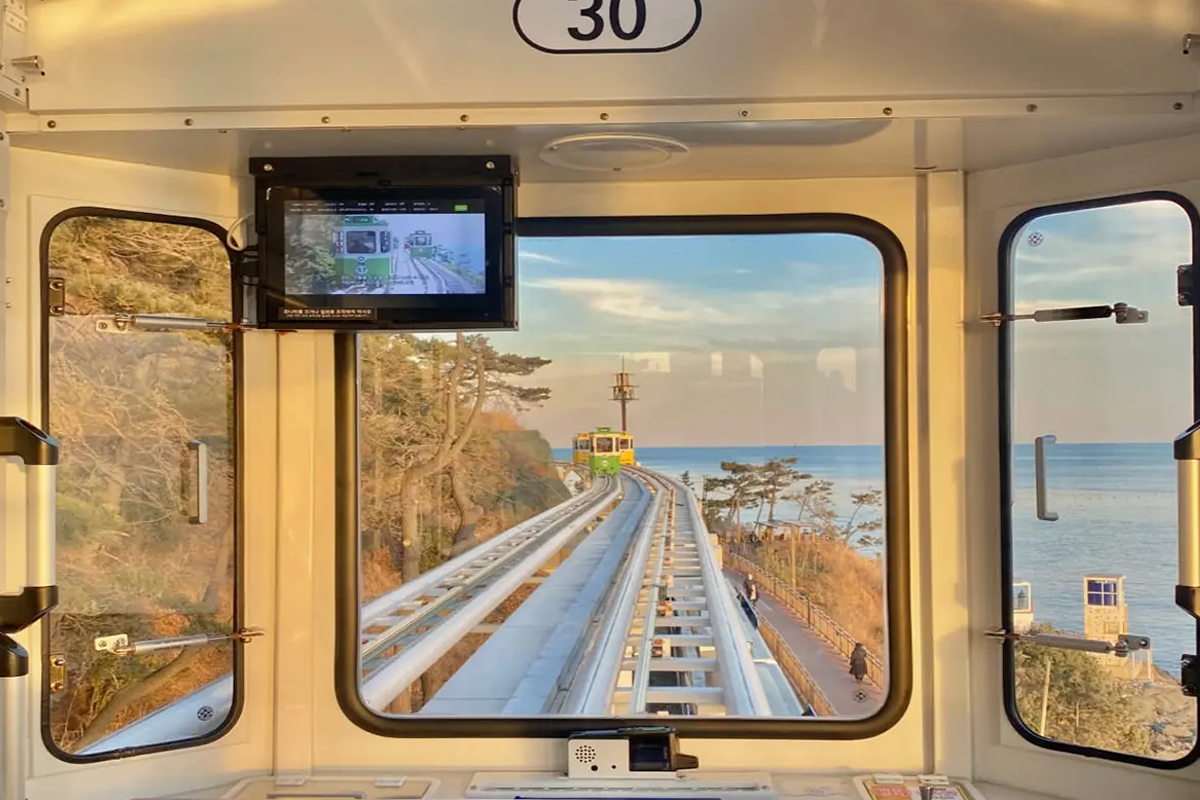 View from inside a Haewundae Capsule Train showcasing the track following the scenic coastline of Busan, South Korea. The interior of the train is visible with the rails stretching towards the horizon and the sea glistening under the sunlight.