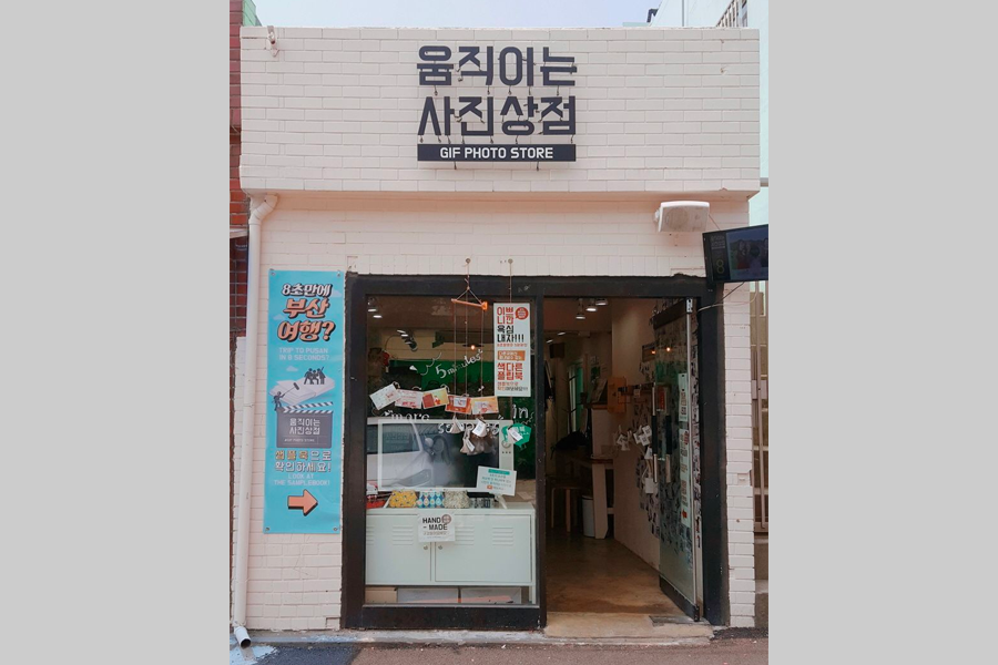 한국 부산 감천문화마을에 위치한 움직이는 사진 상점의 외부 모습. 흰색 벽돌 외관과 다양한 안내 문구가 걸려 있는 입구가 보인다.
