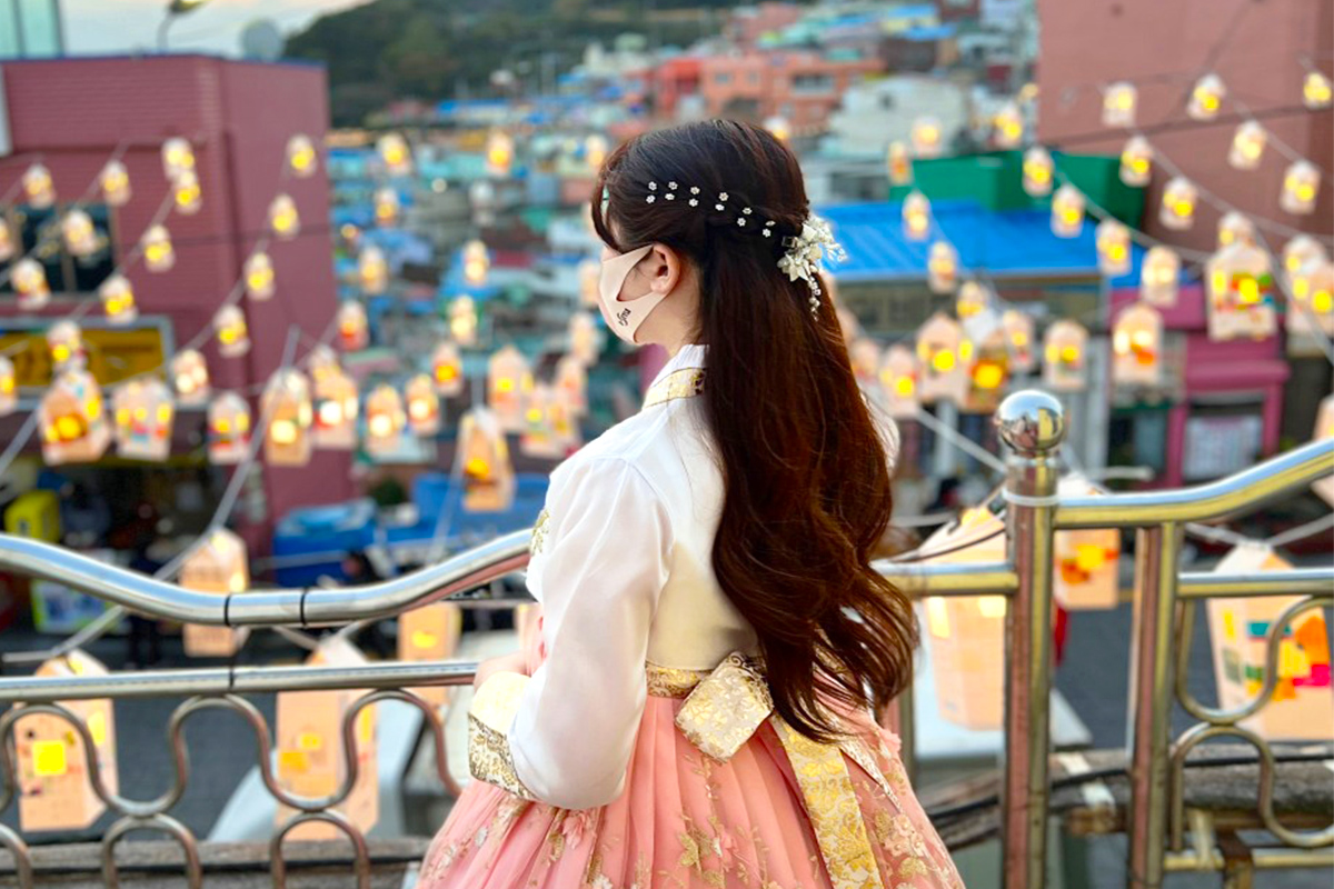 Người phụ nữ mặc hanbok truyền thống, đứng trên ban công nhìn ra một khu phố đầy đèn lồng tỏa sáng, tại Làng Văn hóa Gamcheon, Busan.