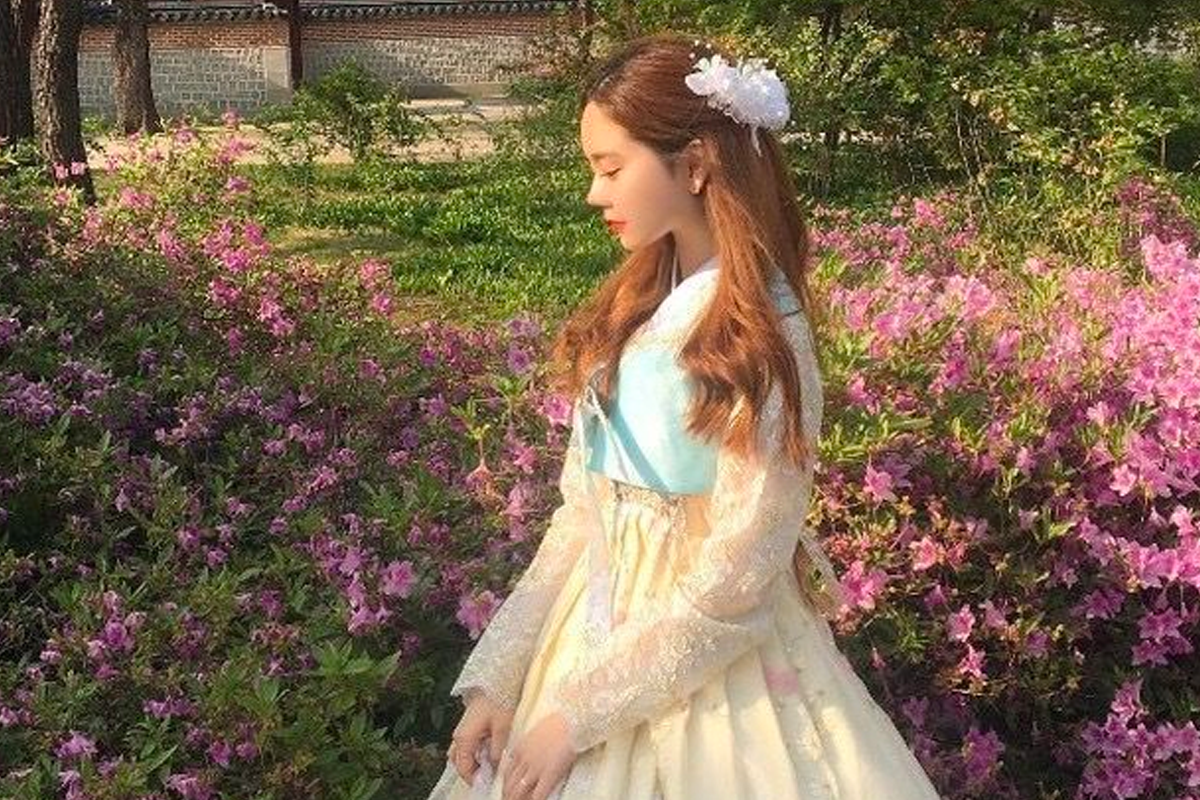 展示一名女子穿著淡藍色韓服站在花園裡，背後是一片花的背景。」