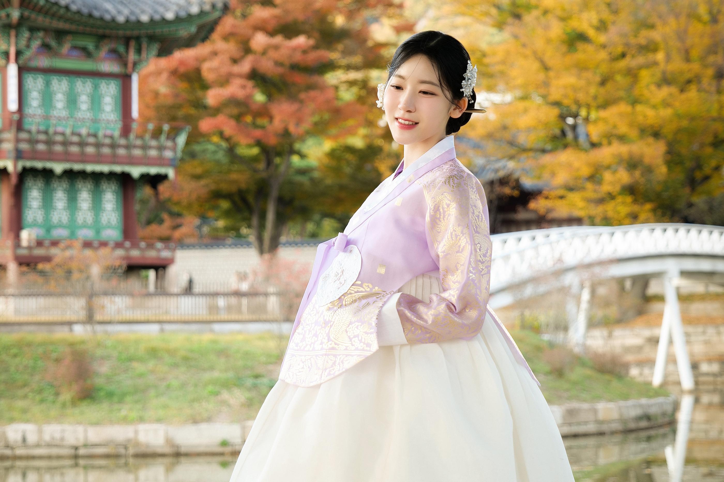 韓国の伝統的な景色を背景に、淡い紫色の韓服を着た女性が立っている。
