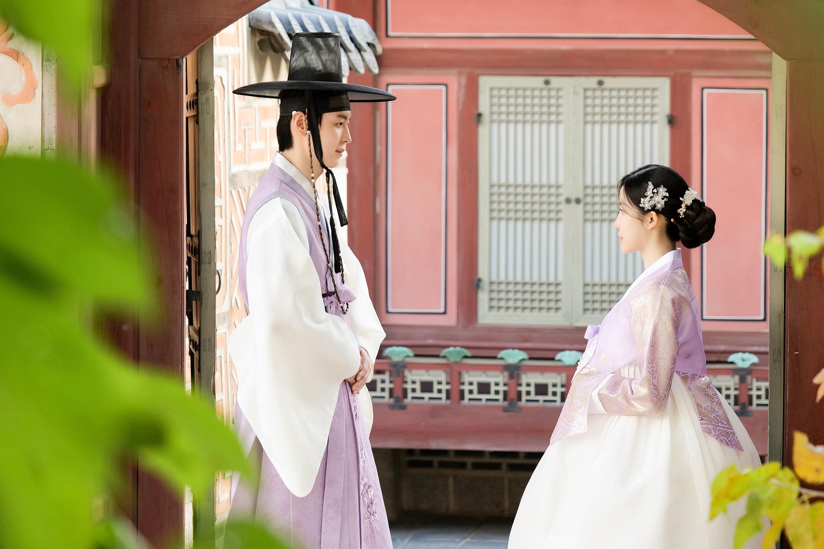 韓国の伝統的な風景を背景に、淡い紫と白のチマチョゴリを着た男女が並んで立っている。