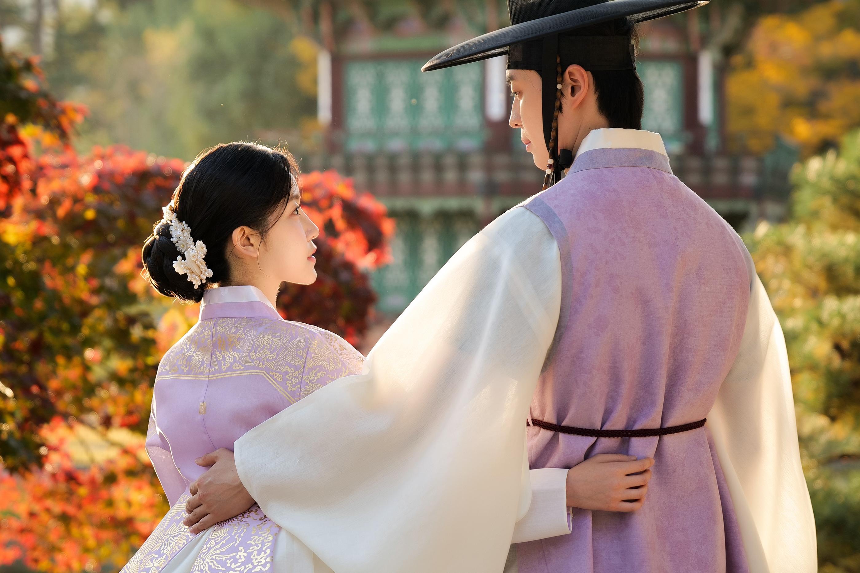 伝統的な韓国の背景を持つ場所で、淡い紫色の韓服を着たカップルが腕を組んでいるロマンティックなシーン。