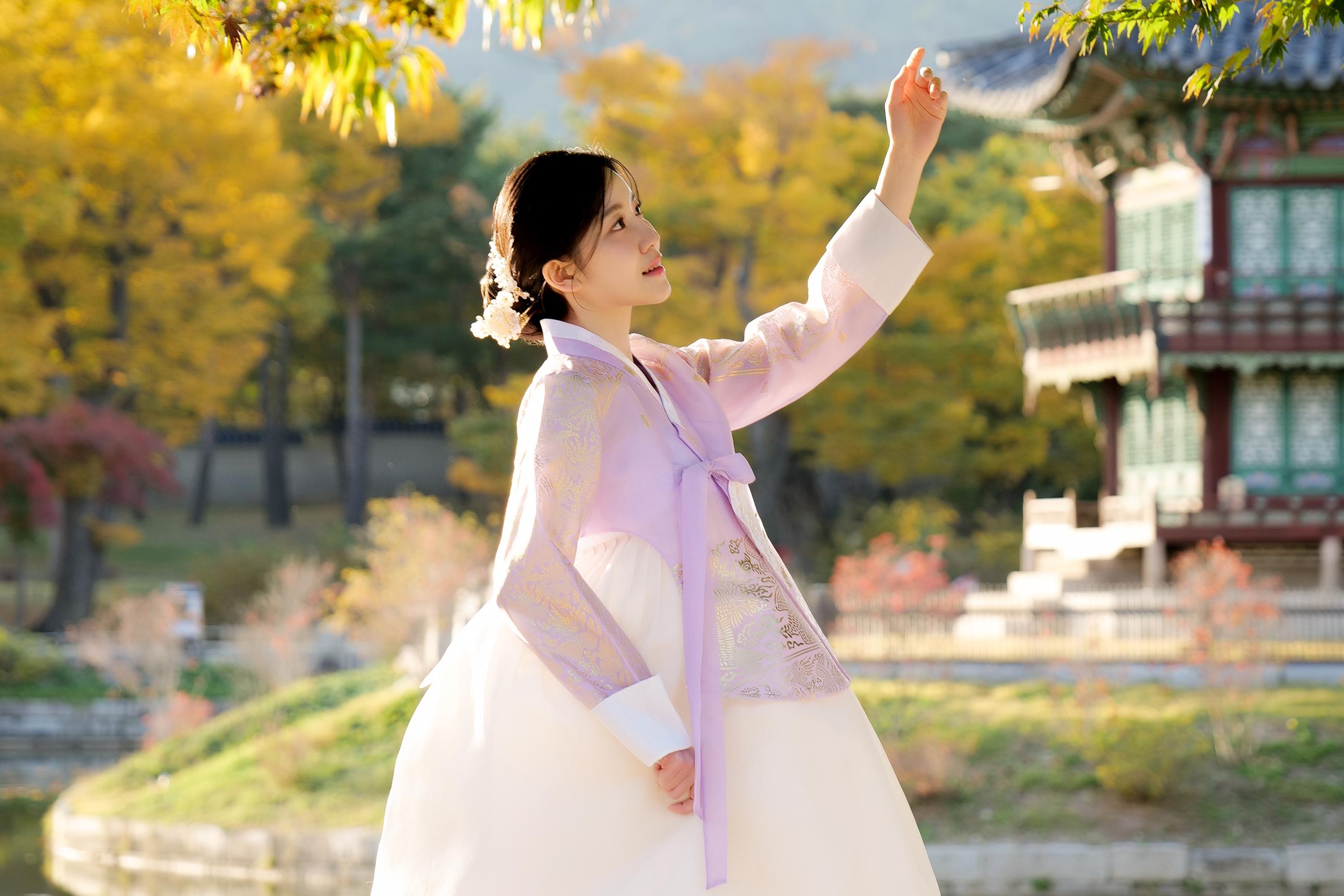 伝統的な韓国の背景に、淡い紫色のチマチョゴリを着た女性が木の下でポーズをとっている。