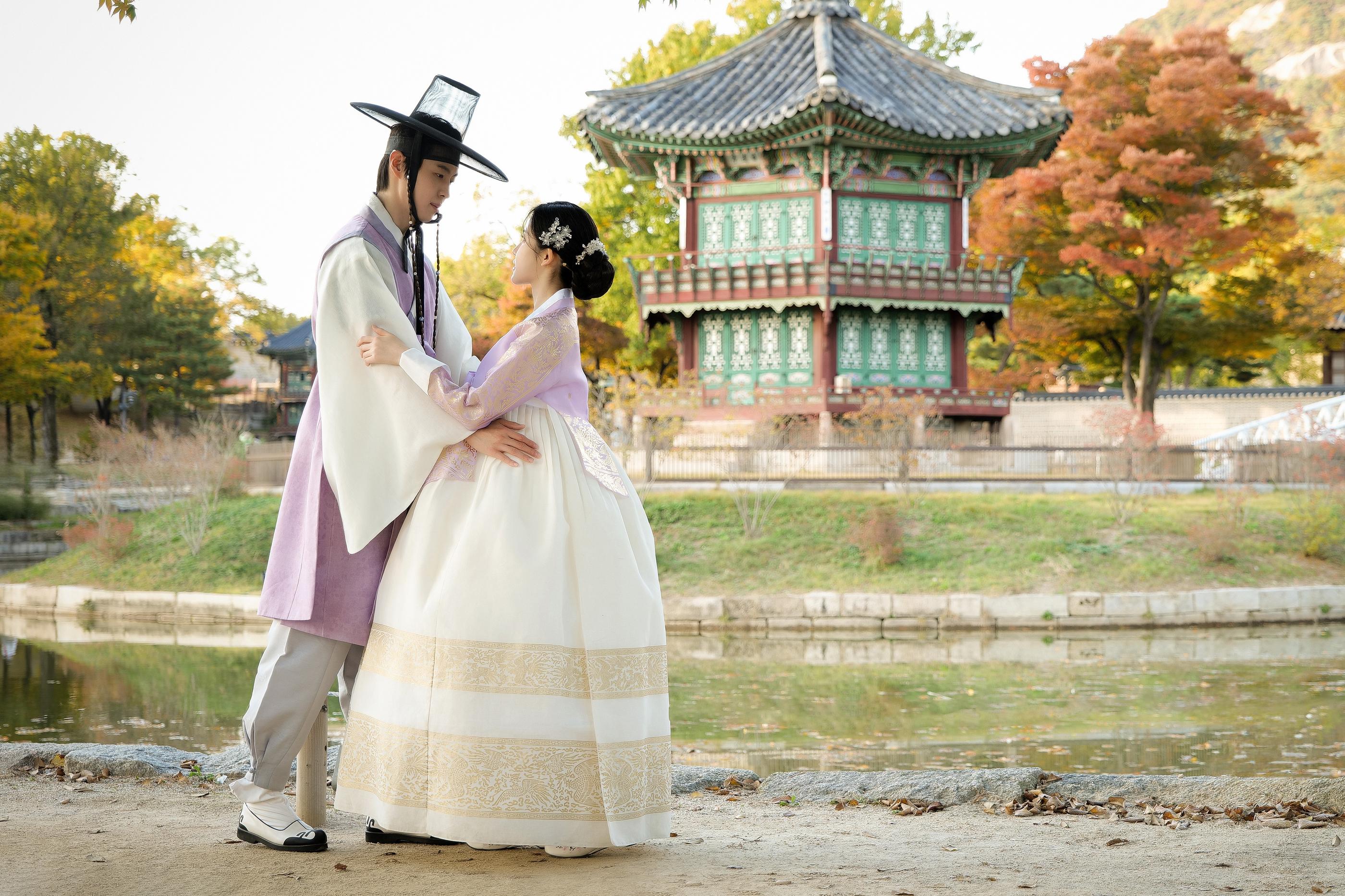 緑の塔を背景にしたカップルの写真。男性は淡い紫の韓服を着ており女性は同系色のチマチョゴリを着た姿。