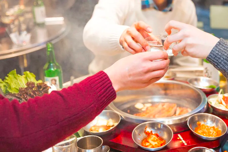 Friends enjoying a Korean barbecue meal, raising glasses for a toast around a sizzling grill.