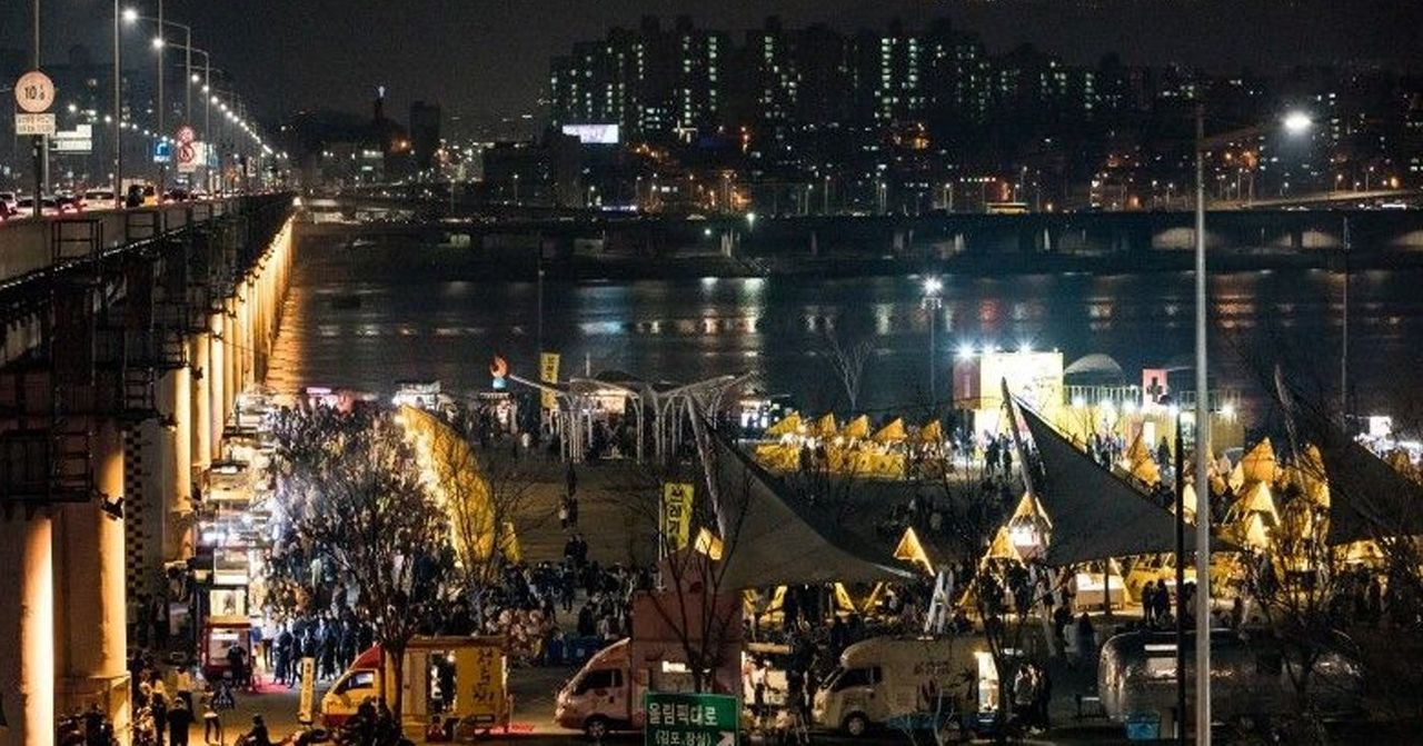 Night view of a bustling Korean night market next to a river in Seoul, with a crowd of people and food stalls.