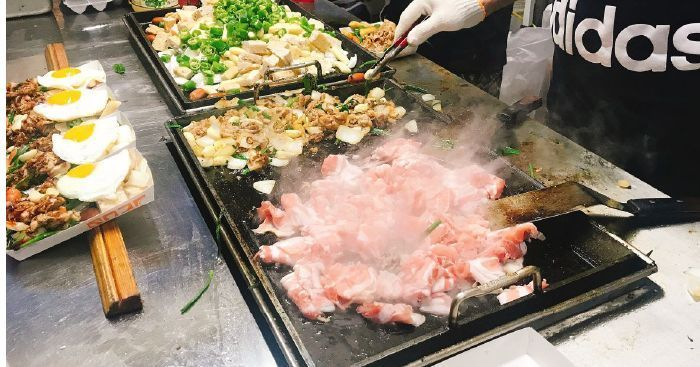Street food being cooked on a flat grill at a Korean market, featuring sizzling meats and vegetables.