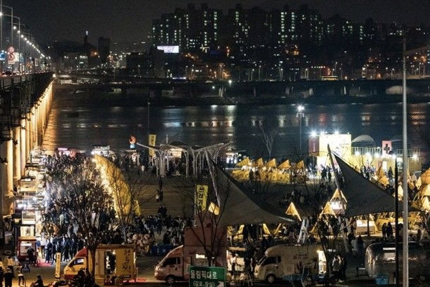 首爾某個夜市的夜景，全景呈現河岸兩旁的燈光和熱鬧的人羣。