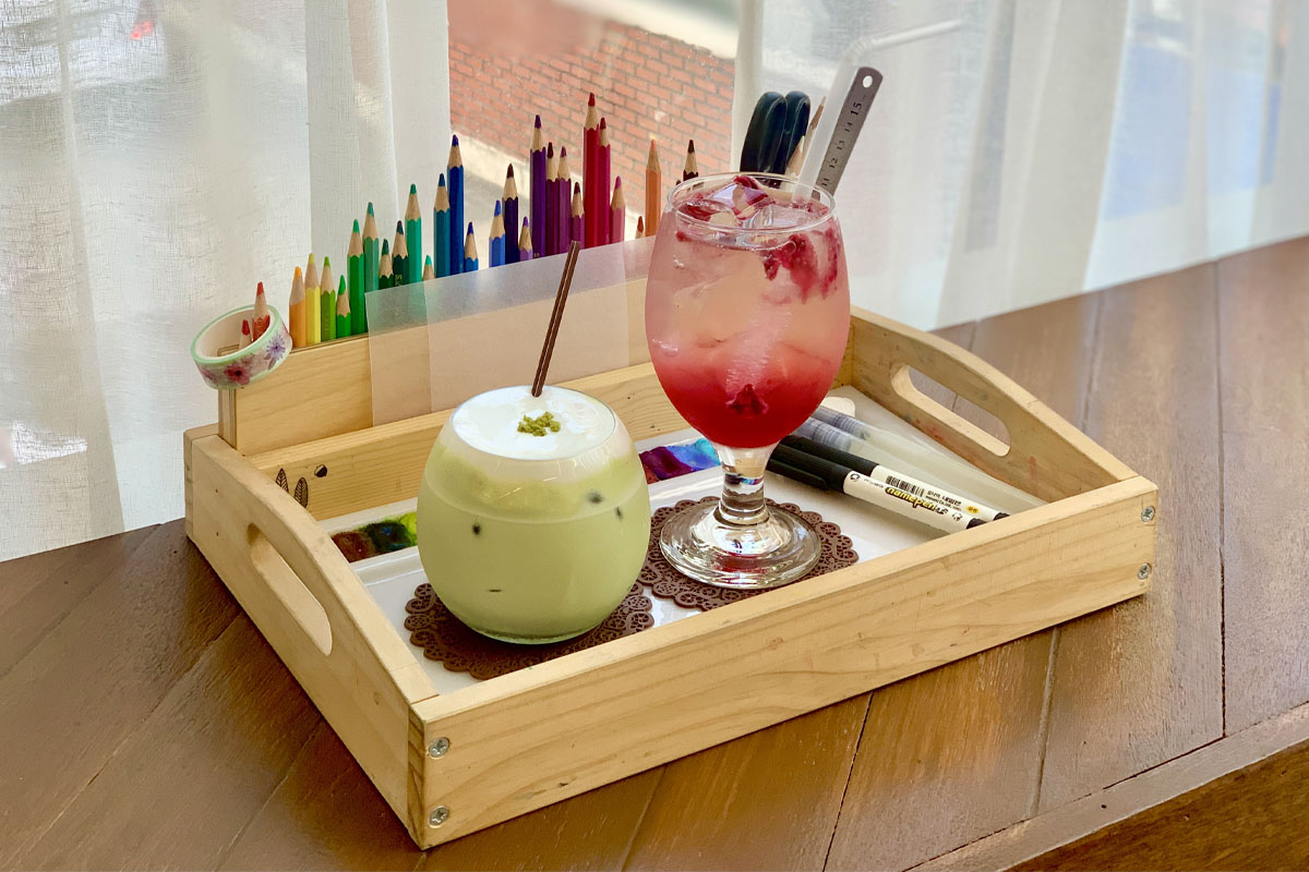 꼬마와 프루츠 색연필, 반짝이는 색감의 컬러폰트가 담긴 음료 트레이에 놓인 두 잔의 음료, 한 잔은 우유 크림이 가득 얹어진 푸른색 녹차 음료, 다른 잔은 화사한 핑크빛 에이드, 카페 창가에 놓인 나무 트레이와 컬러풀한 문구들.