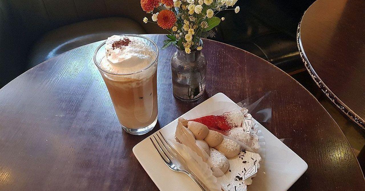 一家咖啡廳的桌上擺著一杯奶油咖啡和一份美味蛋糕，浪漫書香氛圍中點綴著花卉裝飾。